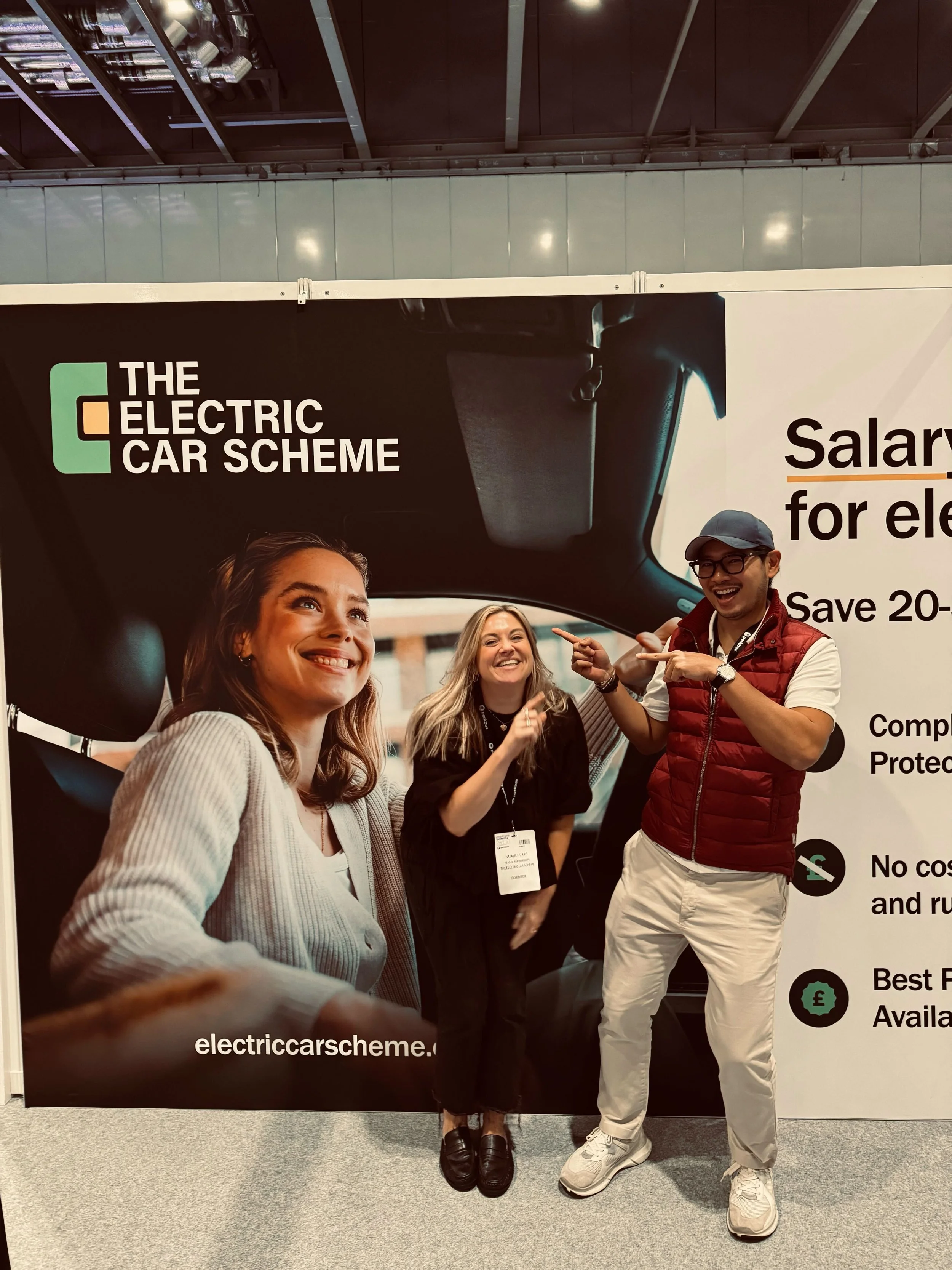 People at an exhibition standing in front of a large sign that reads 'The Electric Car Scheme'. The sign features images of a woman smiling and a man pointing, with text about electric cars, salaries, costs, and availability.