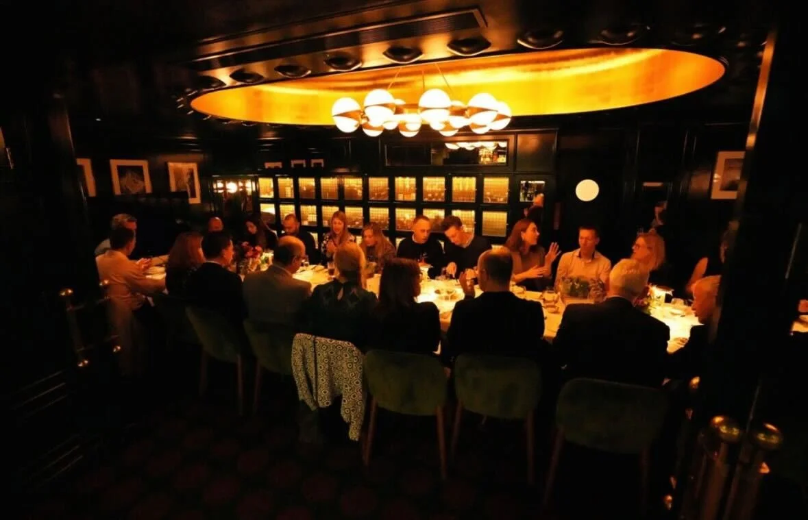 People gathered for a dinner meeting at a long table in a dimly lit, upscale restaurant with modern lighting and dark decor.