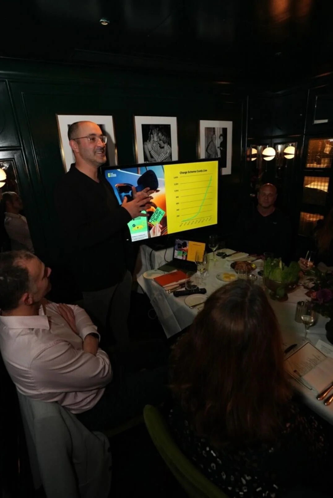 A man is giving a presentation at a dinner event, standing next to a large screen displaying a graph titled 'Charge Scheme Card Line.' Several people are seated around the table, listening attentively. The setting appears to be a dimly lit restaurant