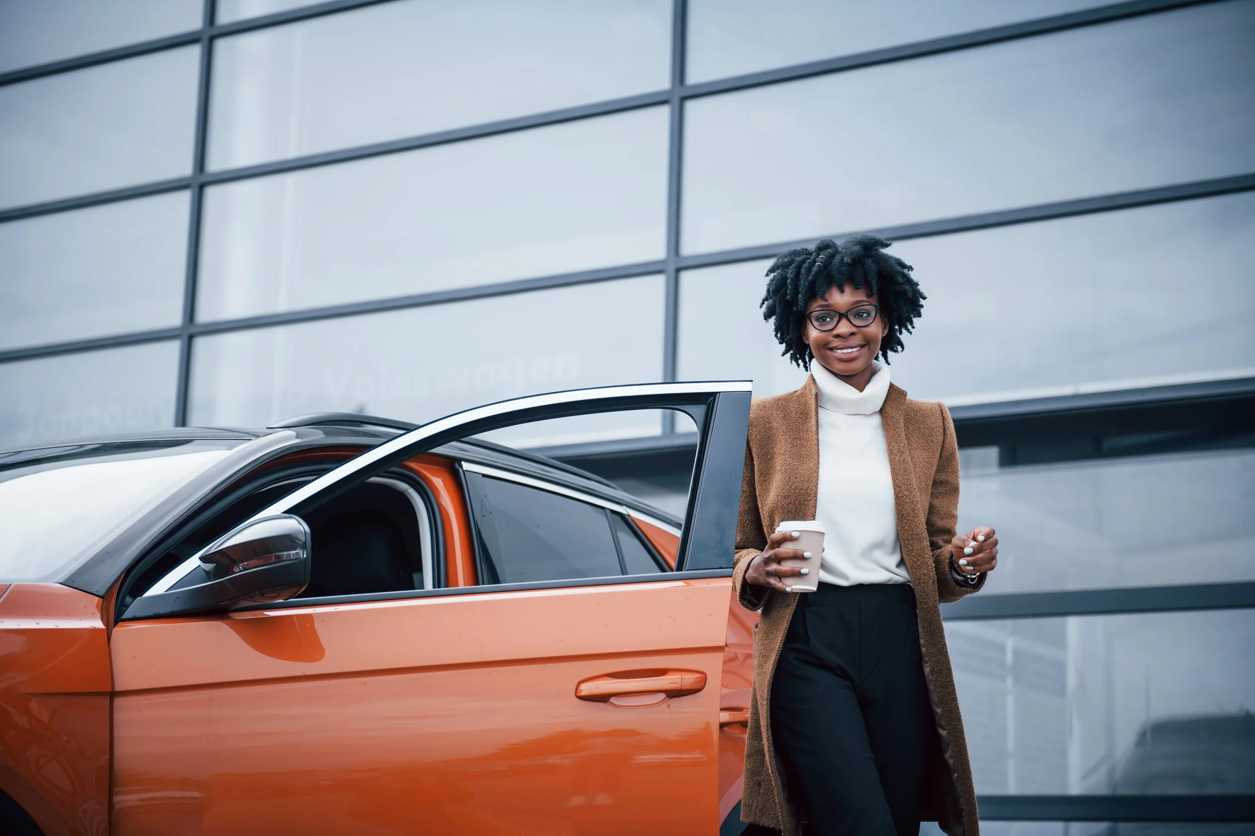 A woman with glasses, curly hair, a white turtleneck, and brown coat smiling while holding a paper coffee cup, standing outside next to an orange and white car in front of a modern building with large glass windows.