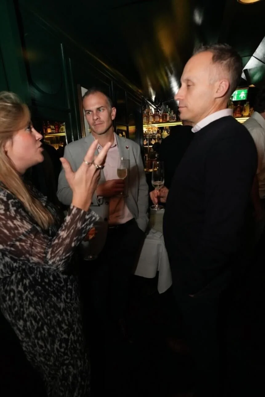 Three people having a conversation at a bar or lounge, with drinks in hand and a background of bottles and ambient lighting.