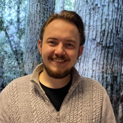 A man with light skin, brown hair, and a beard, smiling in front of a background that resembles a tree trunk with bark.