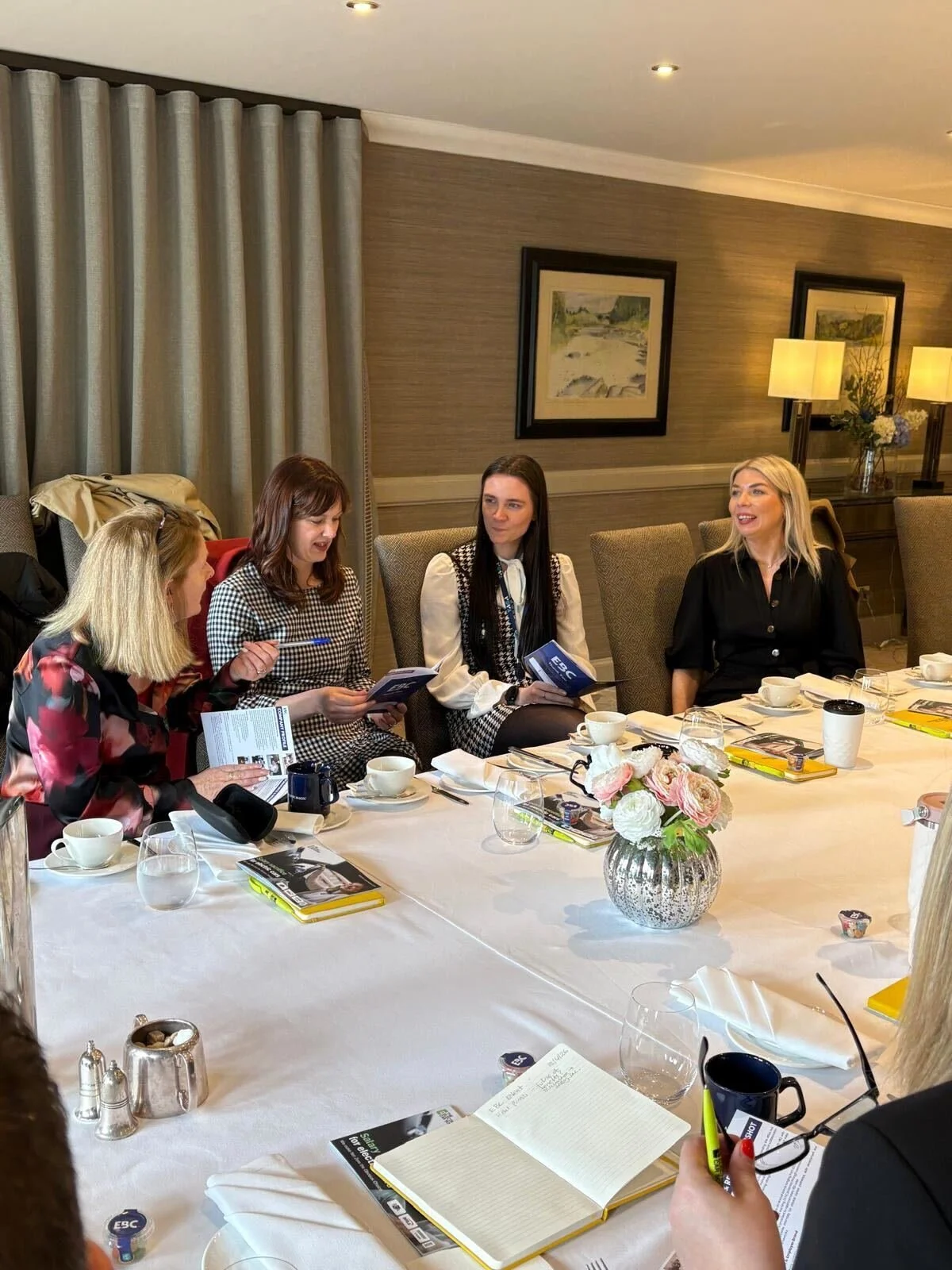 Four women sitting at a conference table in a hotel dining room, engaged in conversation with notebooks and coffee cups in front of them.