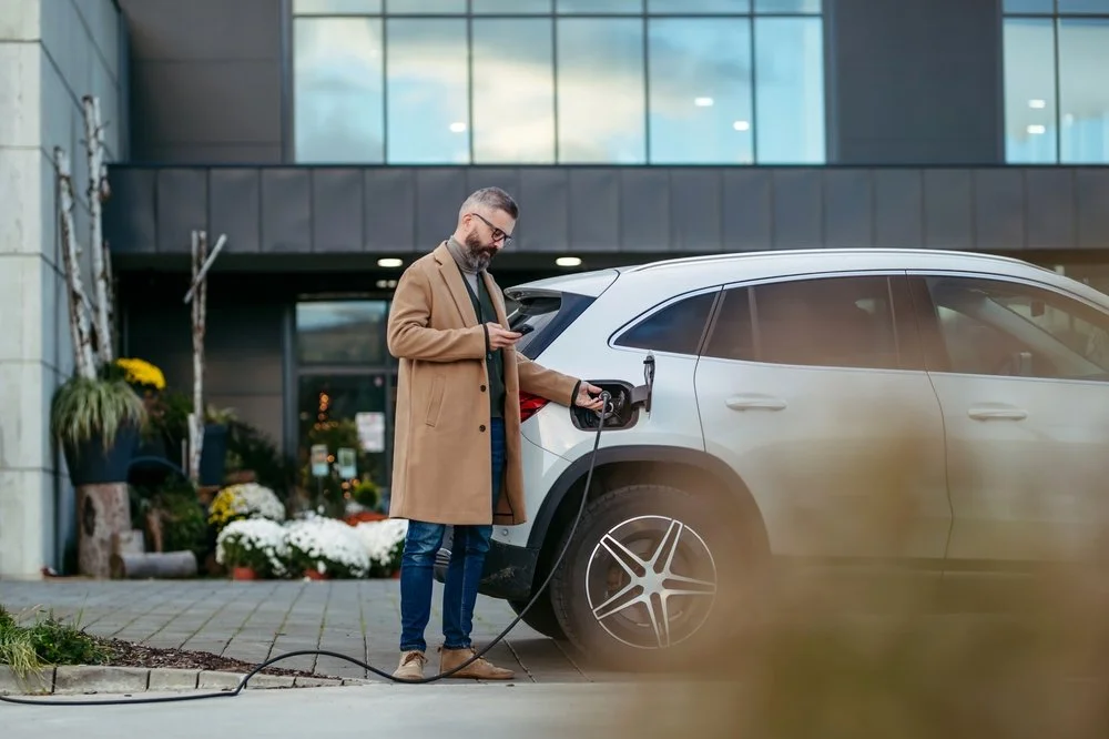 Man plugging in charging plug in electric car before going to shopping. Electric vehicle charging station in front of shop building, supermarket. Charging while shopping.