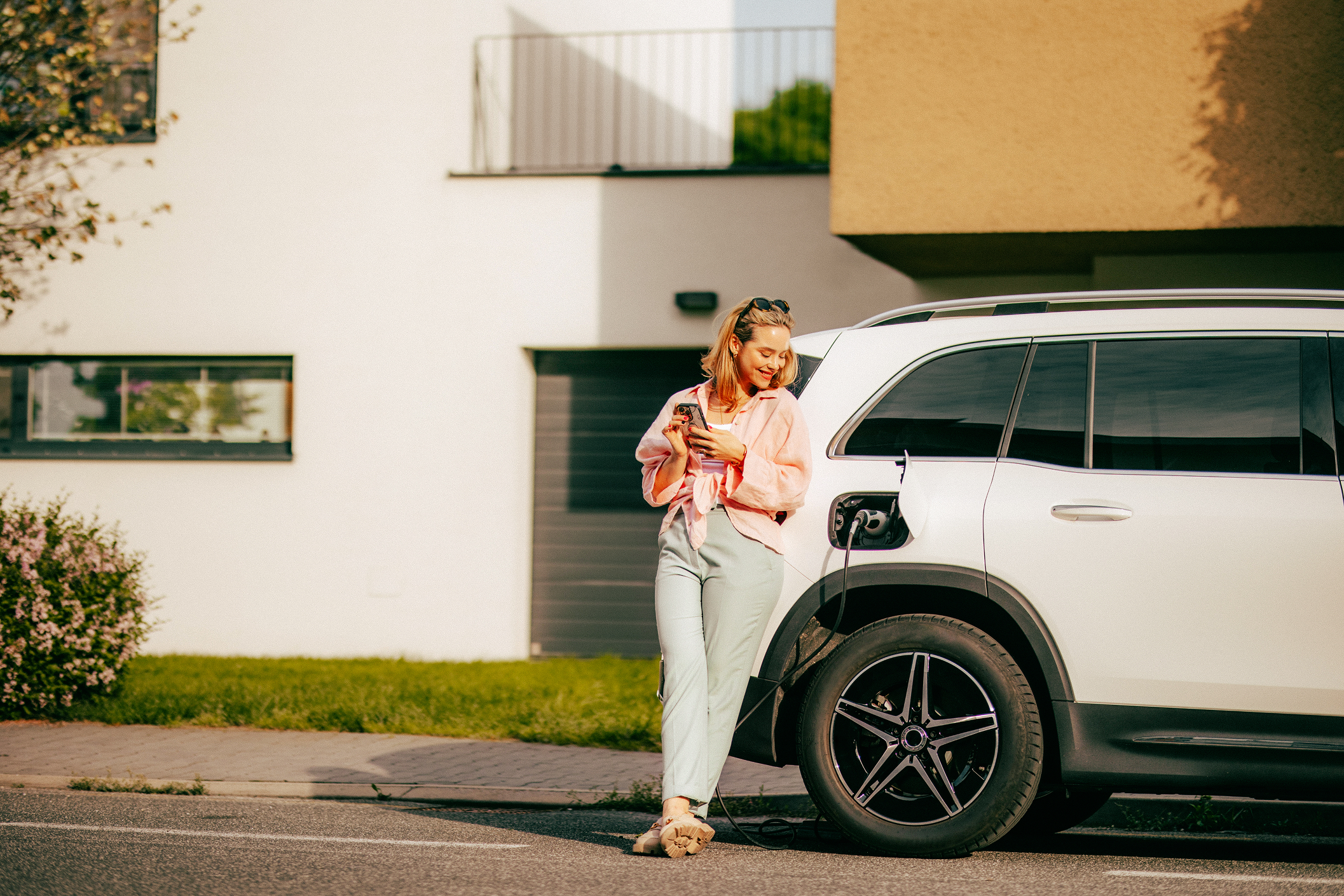A woman with sunglasses on her head, wearing a light pink jacket and white pants, stands beside a white electric vehicle, charging it at a station. She is smiling and looking at her phone.