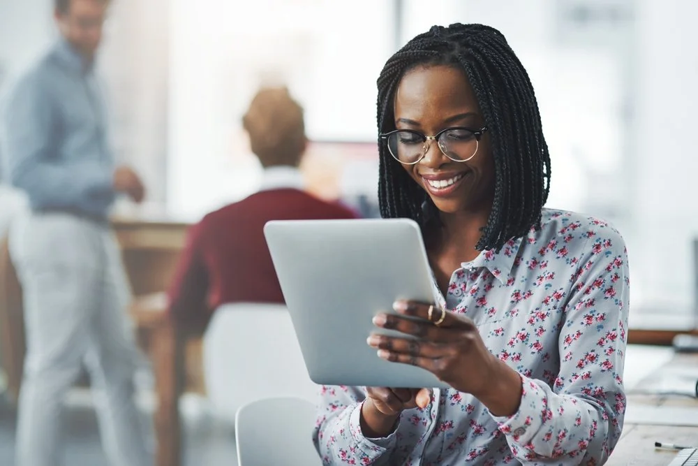 A woman with glasses, smiling and looking at a tablet, in a bright office setting.