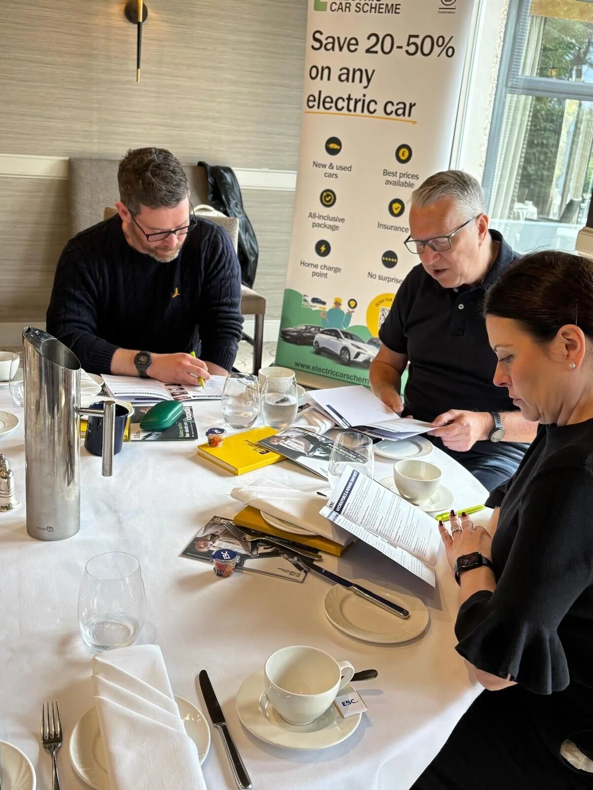Three people sitting at a table during a meeting, looking at documents. A promotional banner in the background advertises a car scheme with discounts on electric cars.