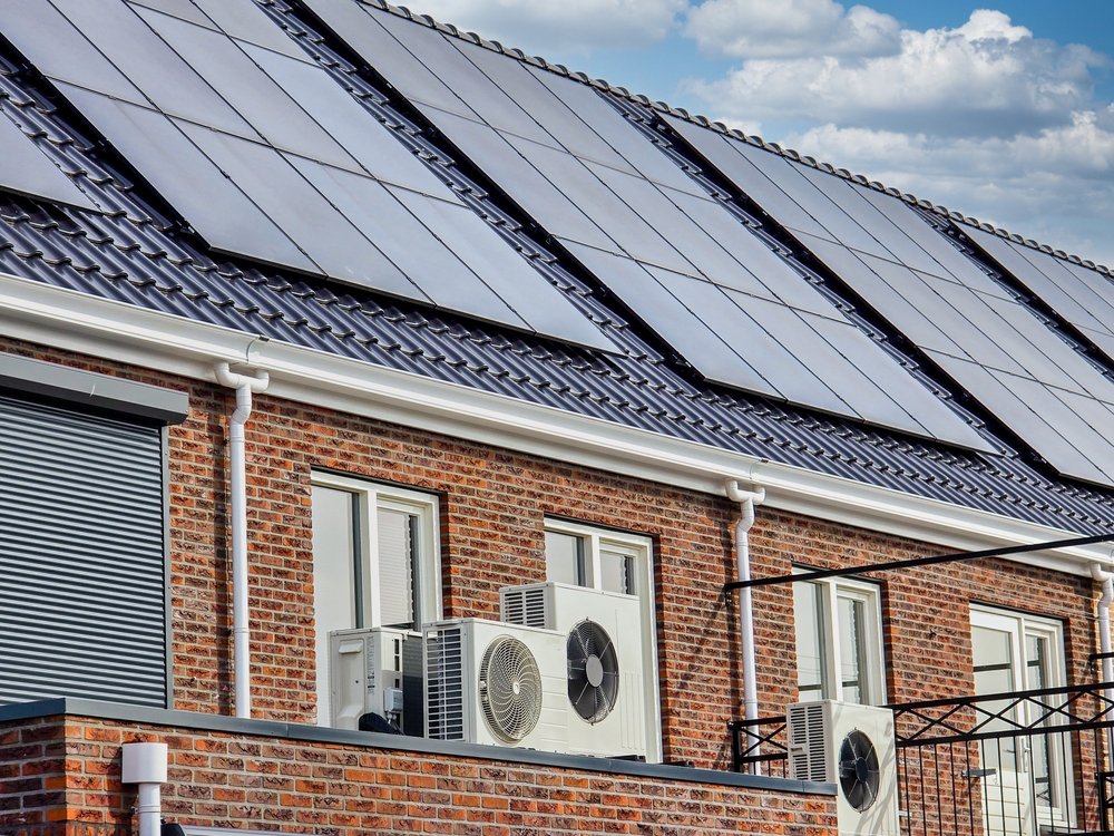 Multiple solar panels installed on the roof of a brick building with air conditioning units and windows.
