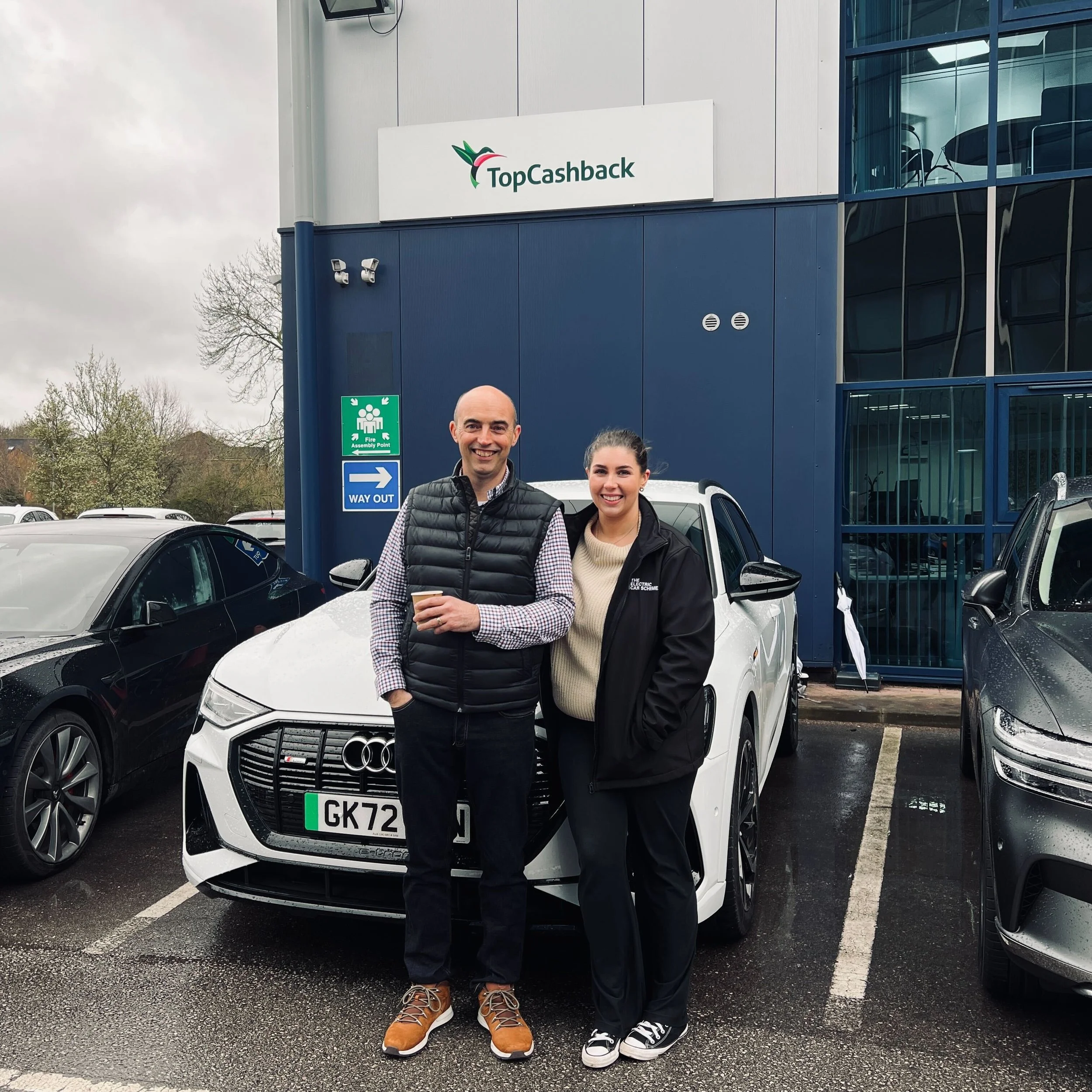 A man and a woman standing together in a parking lot in front of a white Audi car, with a building behind them that has a sign reading 'Top Cashback'. The man is holding a cup and both are smiling at the camera. It is a cloudy day.