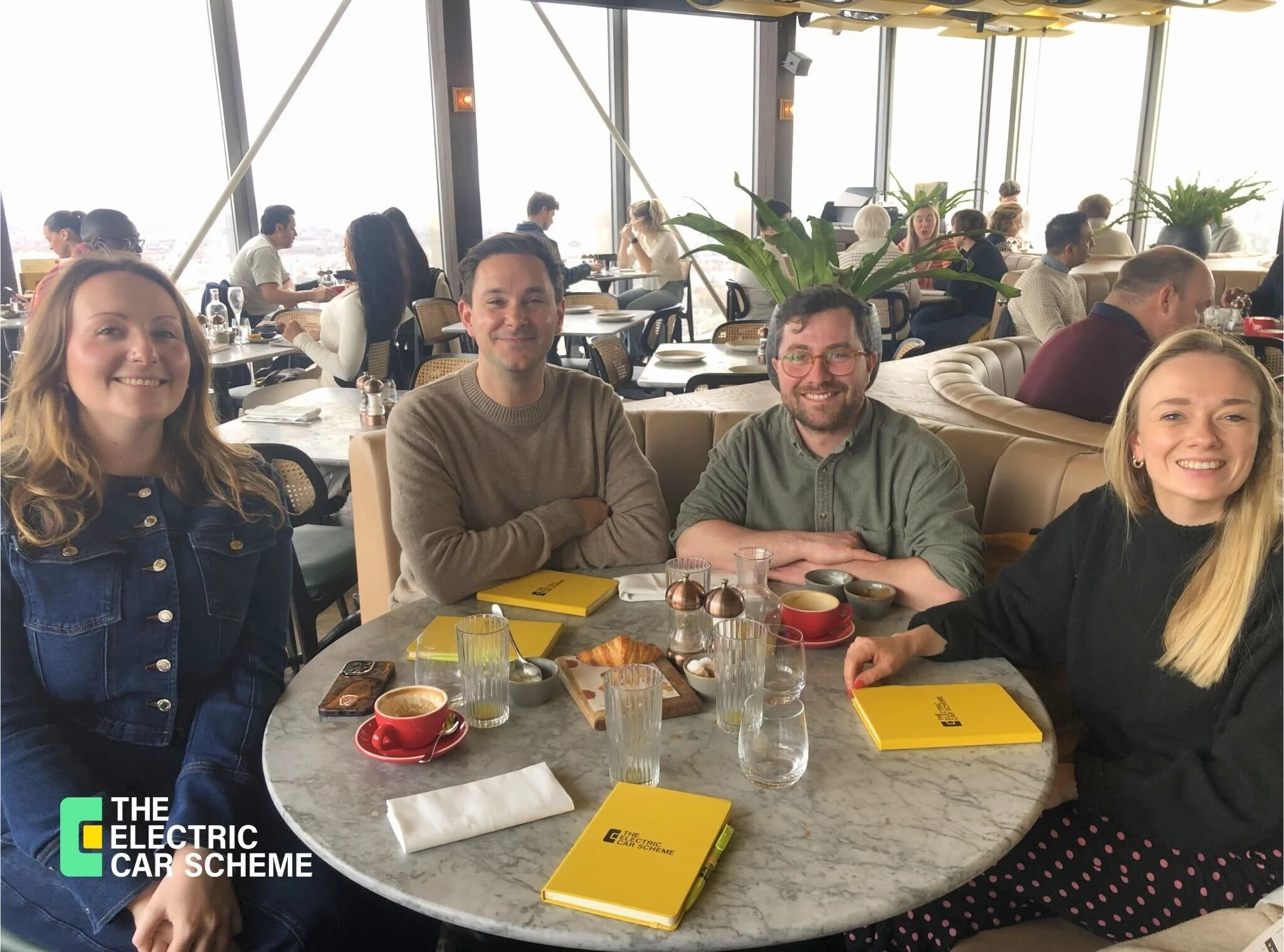 A group of people sitting at a round marble table in a rooftop restaurant with large windows, enjoying breakfast or brunch. There are two women and two men in the foreground, smiling. The table has yellow booklets, coffee mugs, and glasses. In the ba