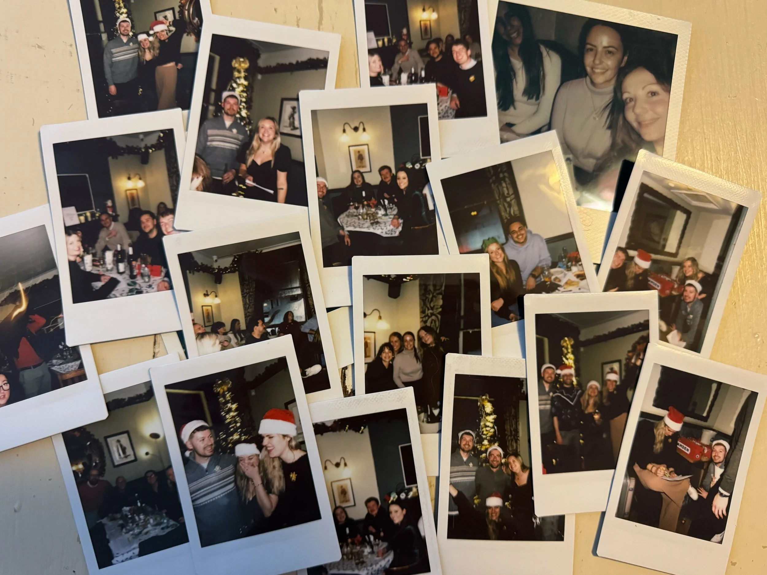 A collection of printed photographs spread out on a table, capturing moments from a holiday gathering with people wearing Christmas hats, smiling, and celebrating indoors with Christmas decorations and festive attire.