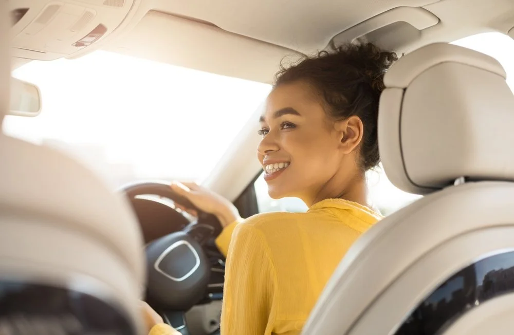 A young woman with curly hair and a yellow top smiling while sitting in the driver's seat of a car.