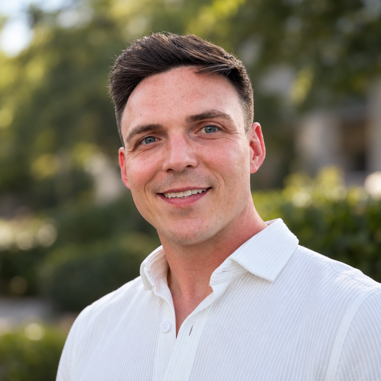 Close-up of a smiling young man with dark hair and blue eyes, wearing a white collared shirt, outdoors with trees and greenery in the background.