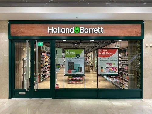 Holland & Barrett store entrance with glass doors, green framing, and promotional posters inside.