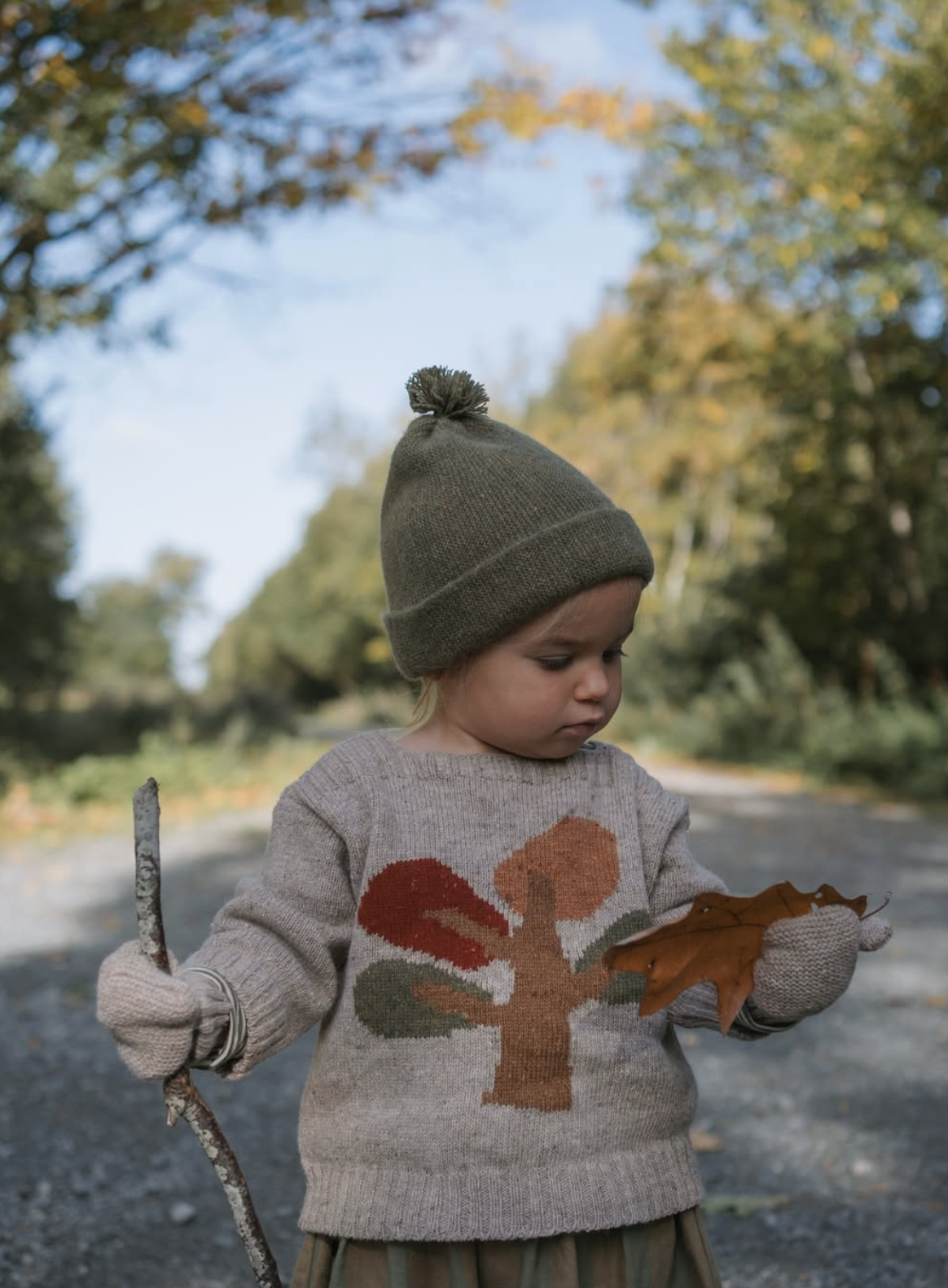 A young child wearing an Organic Zoo knit beanie with a pom-pom, a beige sweater with a colorful tree designed by The Kids Design Consultancy.