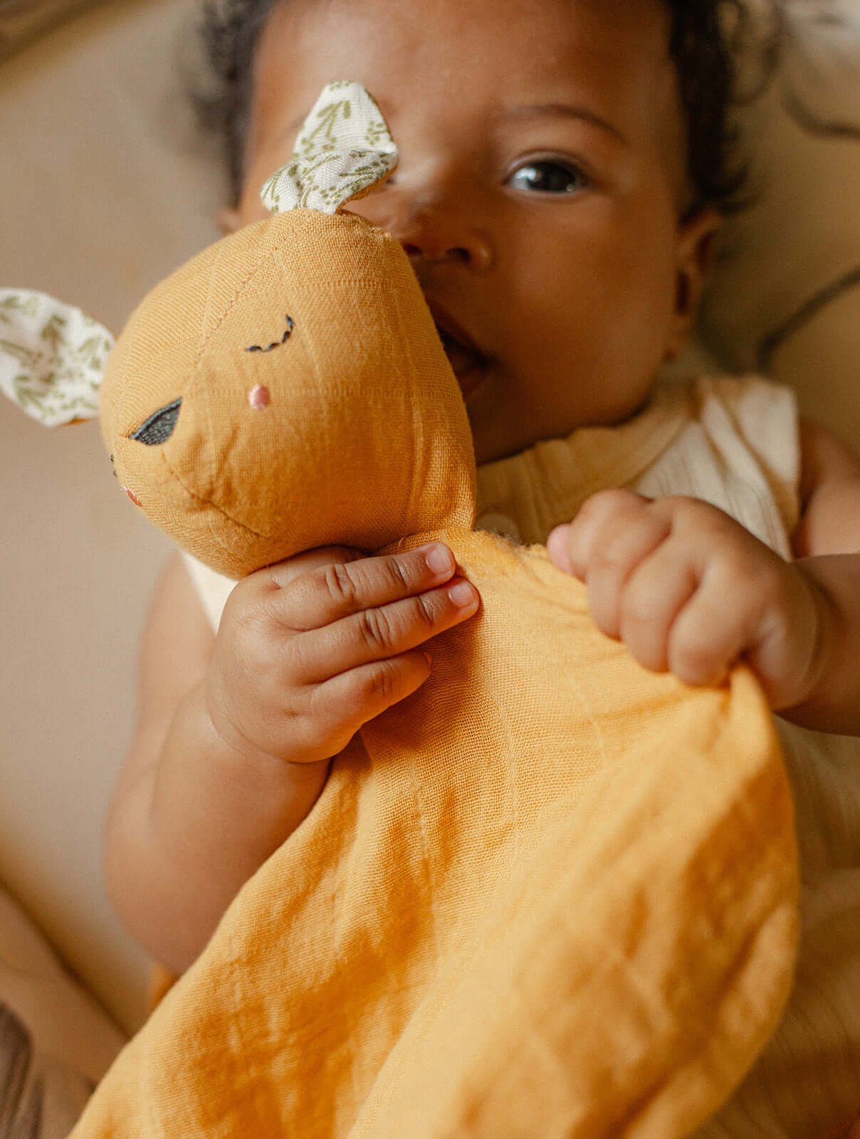 A baby holding a mustard yellow muslin comforter toy.
