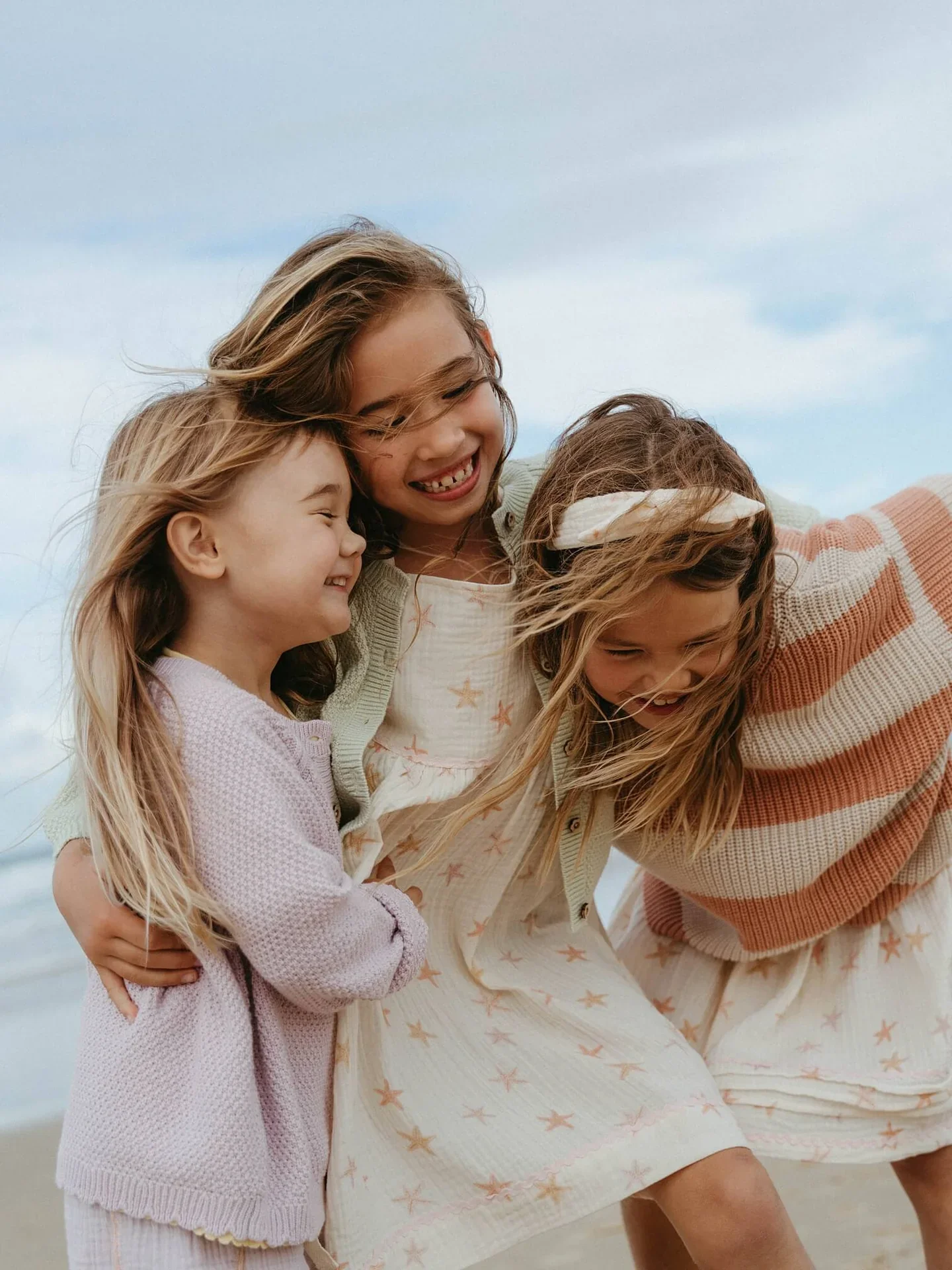 three young girls wearing Yes Mila clothes designed by The Kids Design Consultancy, all smiling and laughing, outside near the ocean during daytime.