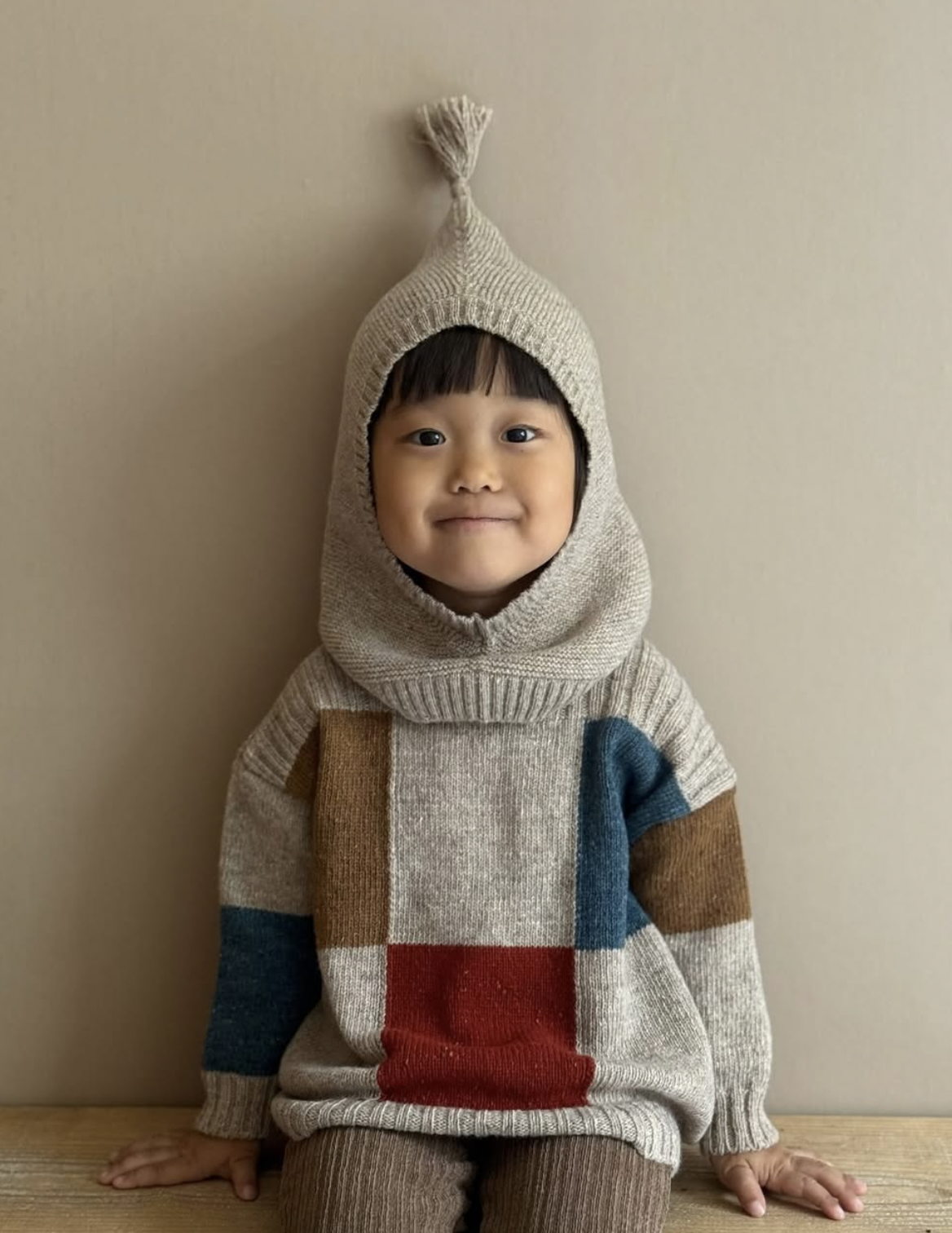 A young child with short dark hair and a smile, sitting on a wooden surface against a plain beige wall, wearing a beige knitted hoodie with a tassel on top and a colorful patchwork sweater.