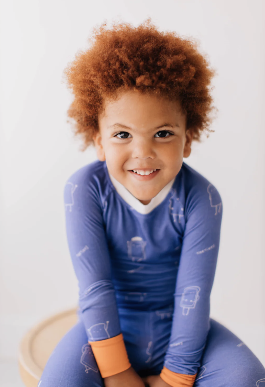 A young child with curly red hair smiling and sitting on a wooden stool, wearing a blue long-sleeved shirt with patterns and orange cuffs, against a plain white background.
