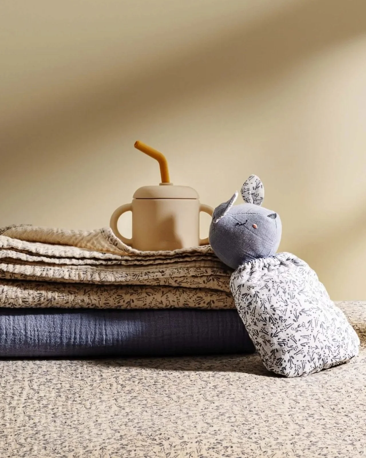 A bunny toy with long ears, one of which has a black leaf pattern, sits on stacked textured blankets. Behind it, a beige sippy cup with a yellow straw rests on the top blanket. The blankets have various soft fabrics and patterns, placed on a neutral-colored surface with a plain, light-colored wall in the background.