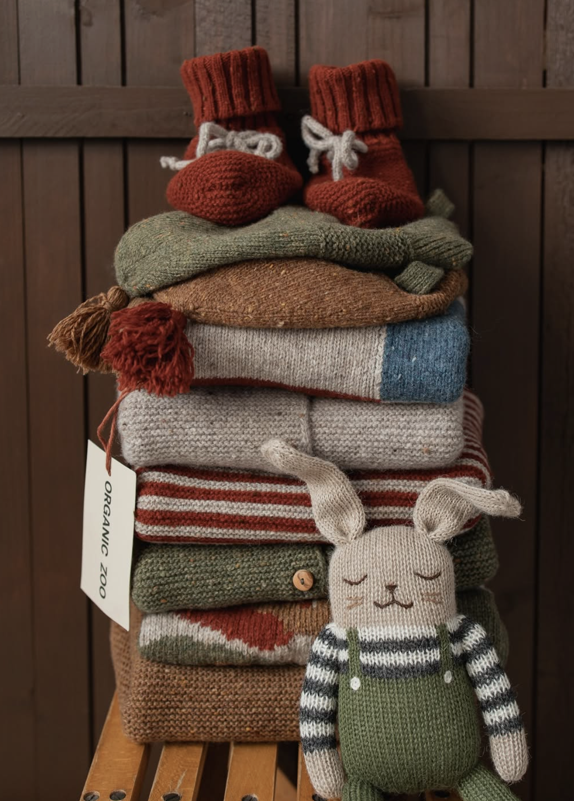 Stacked folded clothes including hats and sweaters beside a plush rabbit toy in front of a wooden background.