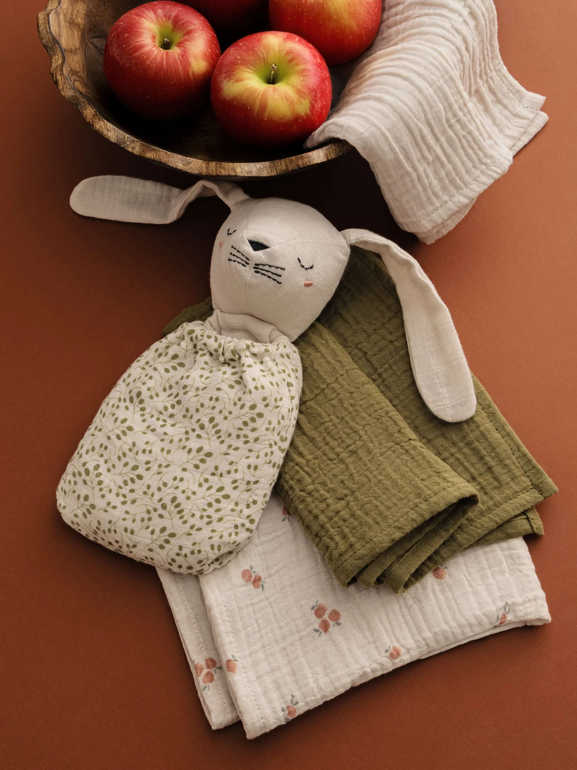 Three red apples in a wooden bowl with a white cloth lining, next to a cloth bunny toy and folded green cloth on an orange surface.