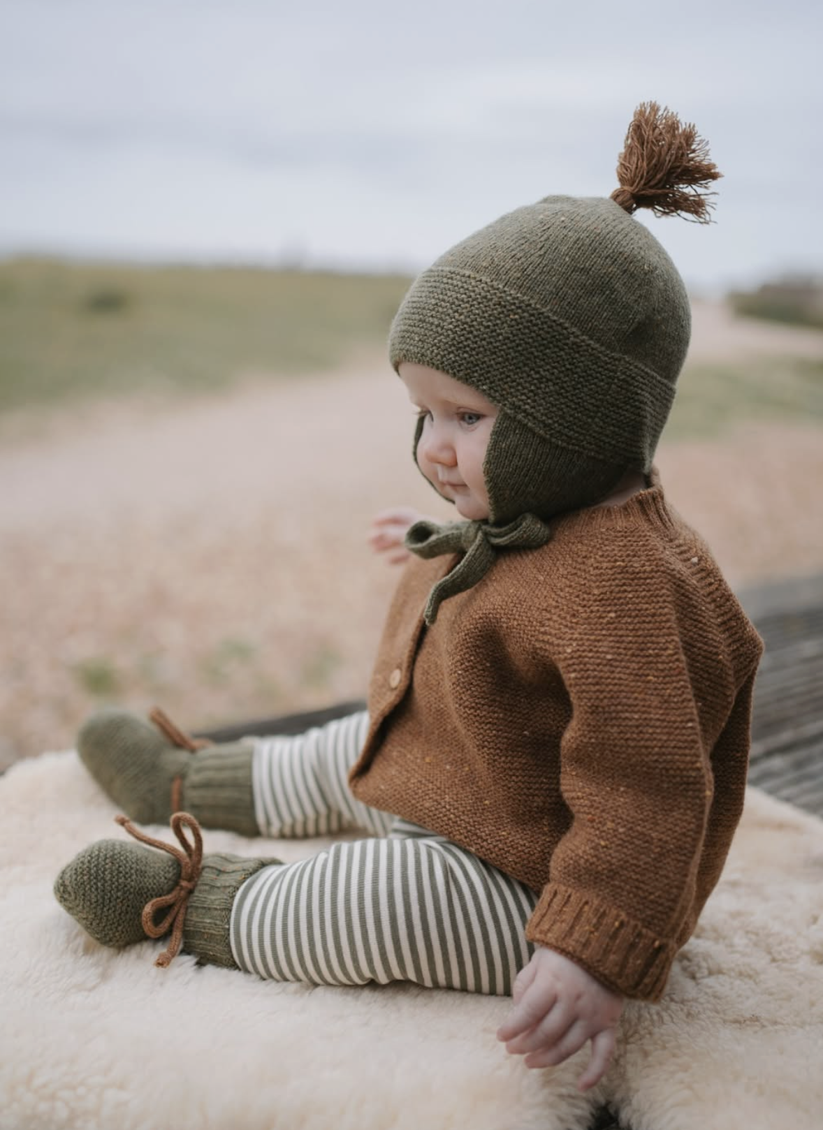 A young child sitting on a soft surface outdoors, wearing a green knit hat with a pom-pom, a brown knit jacket, striped pants, and knitted mittens, with a blurred outdoor background.