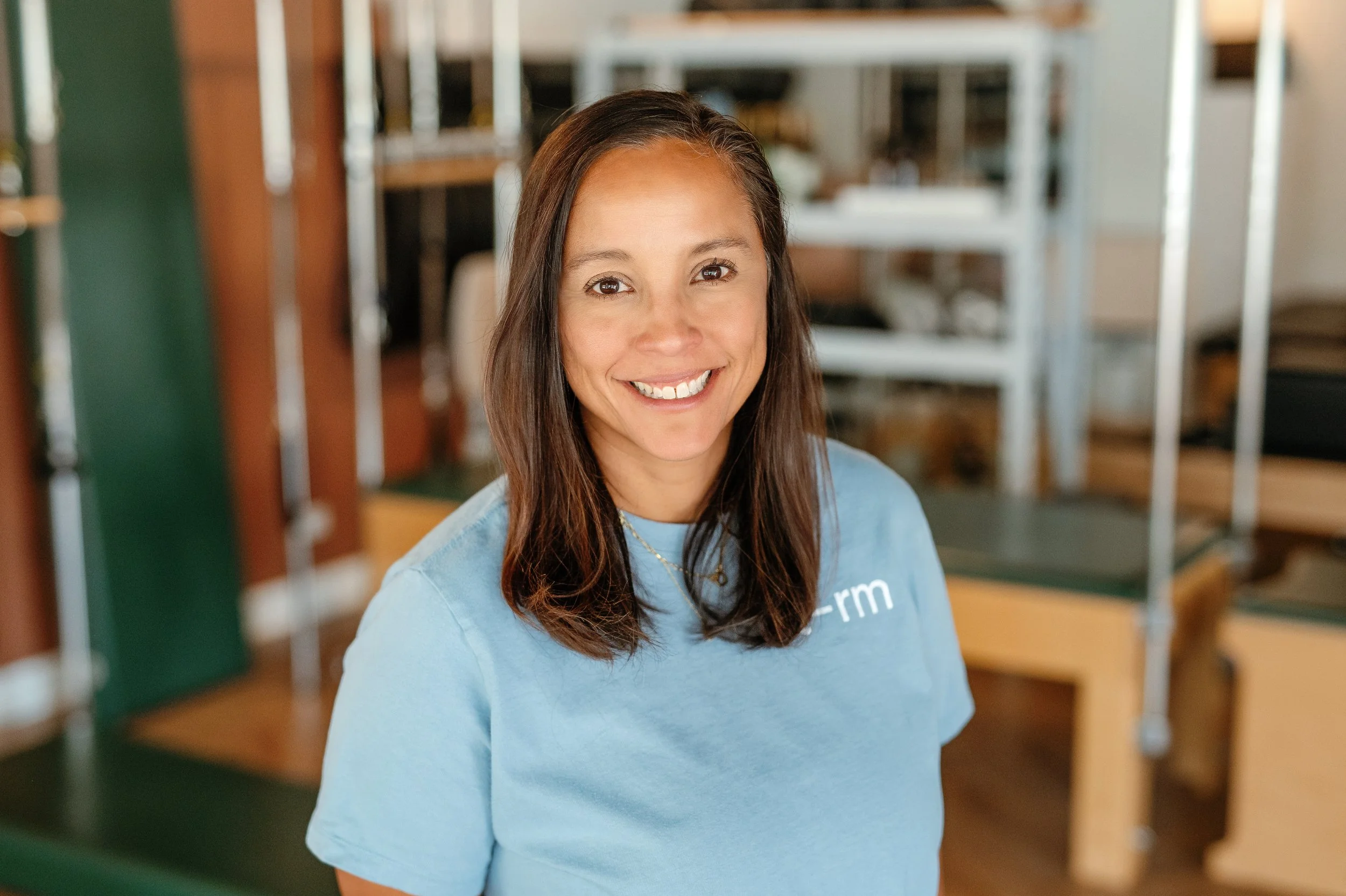 A woman with shoulder-length dark hair smiling while leaning against a wall. She wears a maroon sweatshirt with the logo 'reform PT + Pilates' on it.