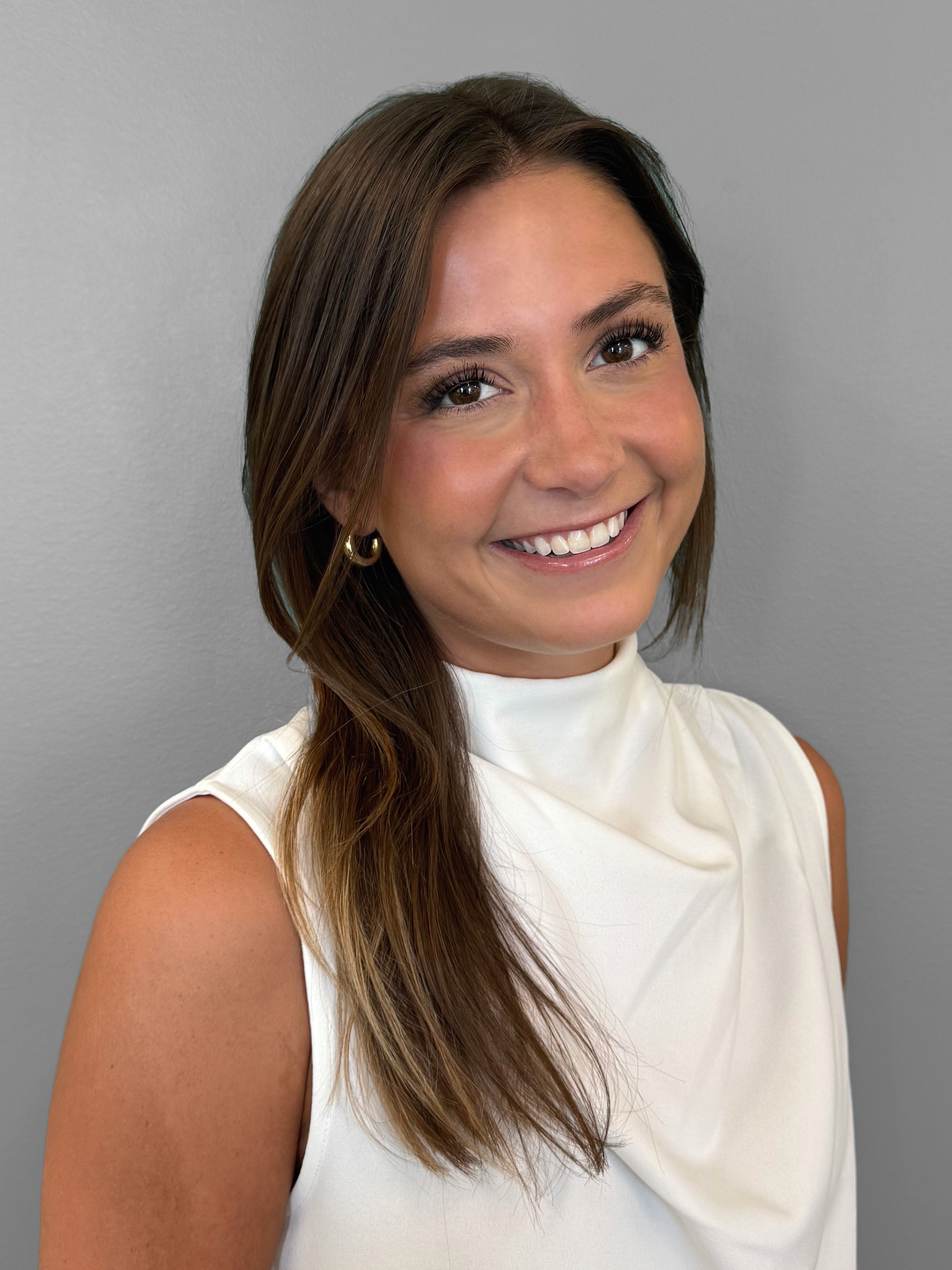 Cayda our MA with long brown hair and light skin, smiling, wearing a white sleeveless top and gold earrings, standing against a plain gray wall.