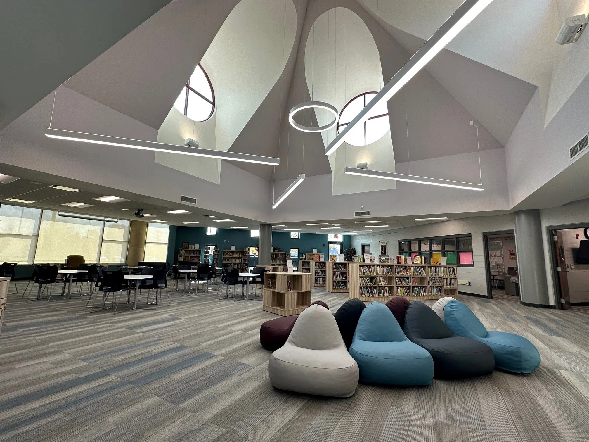 Interior of a modern library with bookshelves, tables, chairs, and bean bag seats near a windowed wall, with a high ceiling and contemporary lighting fixtures.