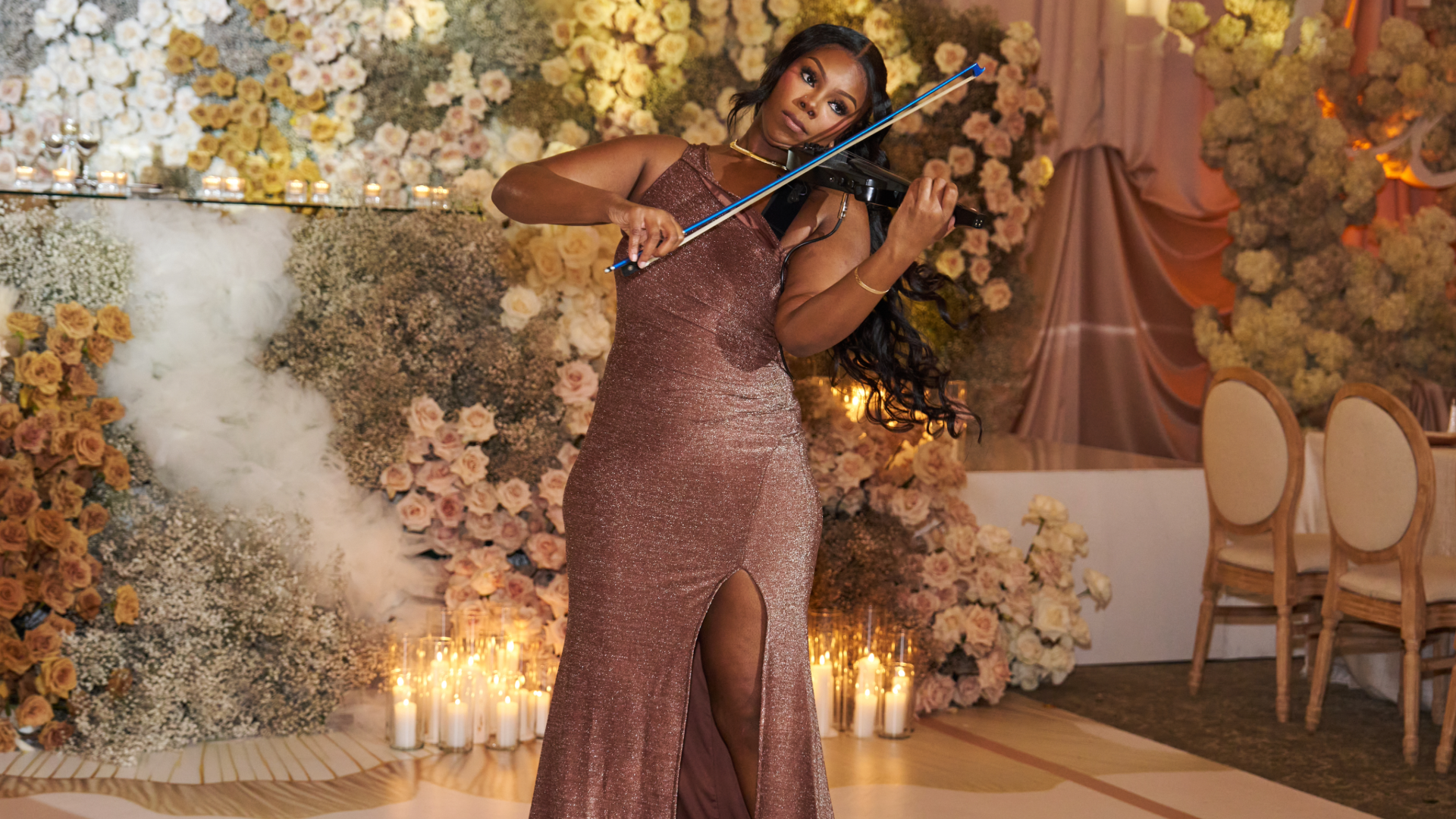 Priya Deberry in a sparkly, mauve gown playing the violin at a floral-themed event with candles in the background.