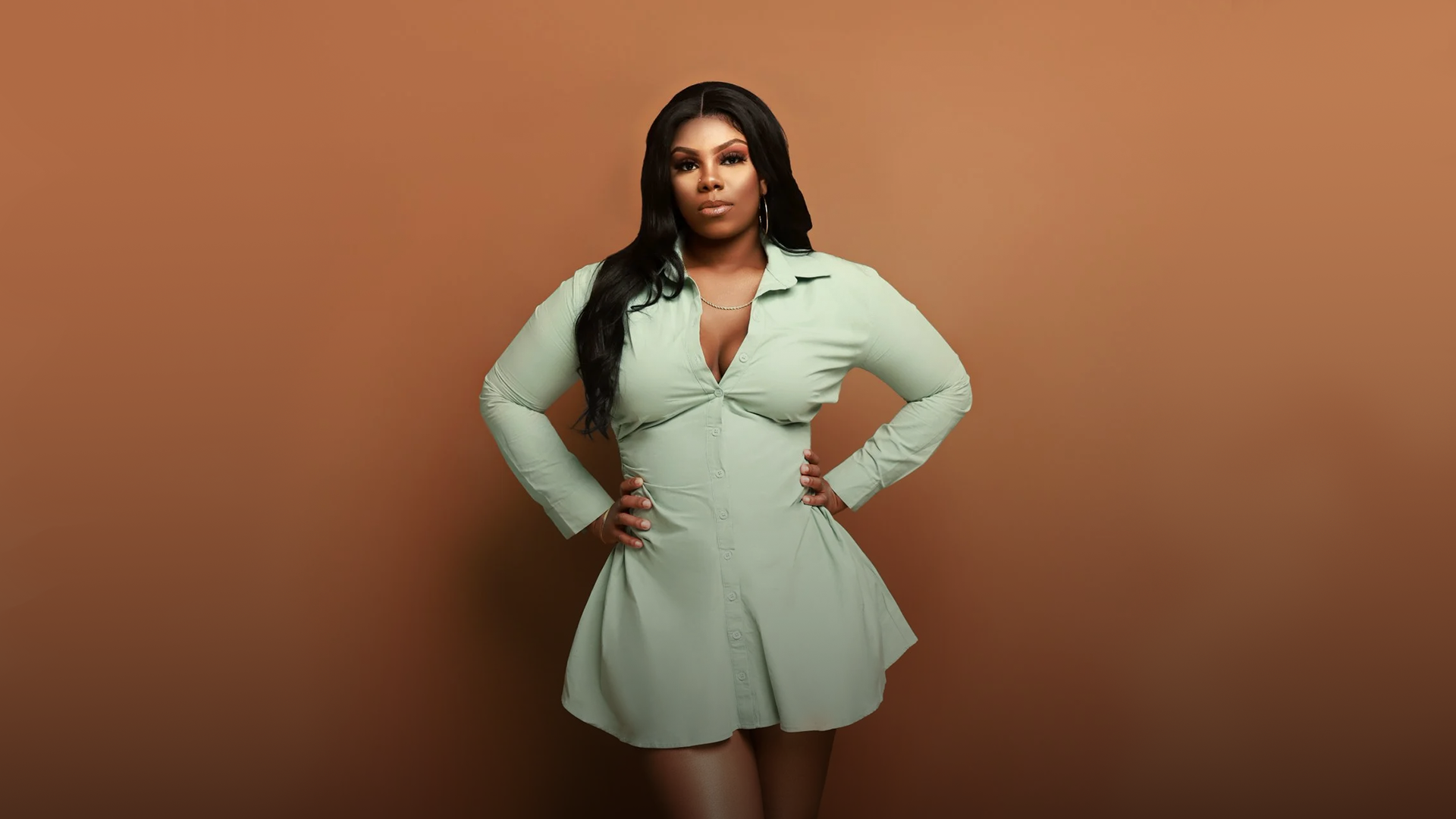 Priya Deberry with long black hair poses with her hands on her hips in front of a brown background. She wears a light green shirt dress with a collar and buttons down the front.