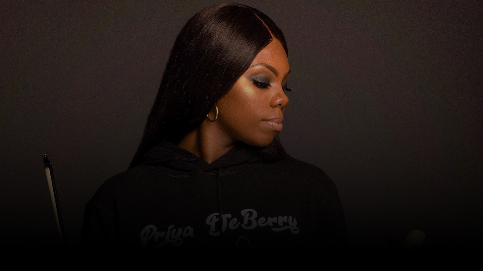 Priya Deberry with dark, straight hair wearing a black hoodie and hoop earrings, looking downwards against a plain dark background.