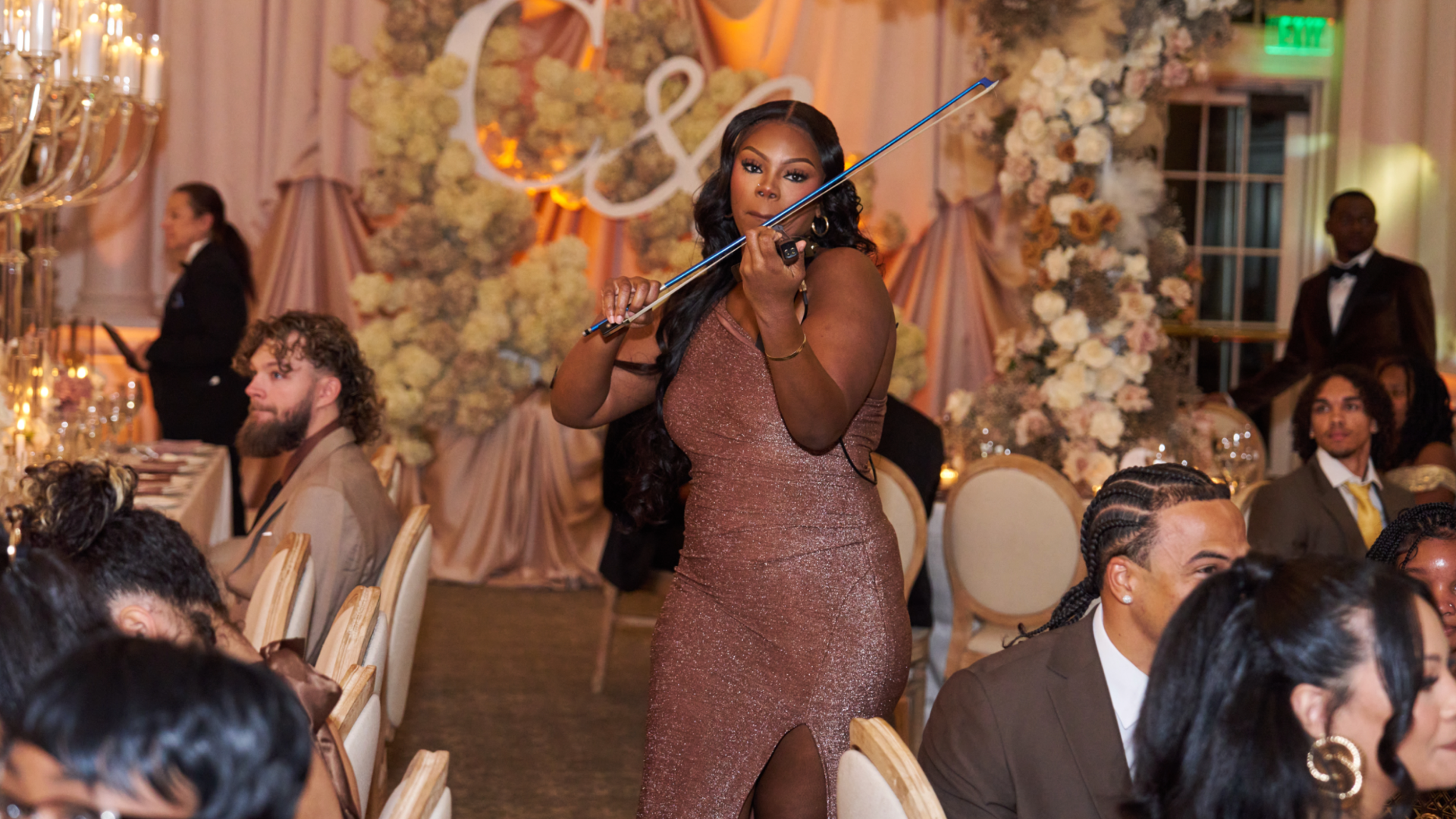 Priya Deberry in a sparkly mauve dress holding a long cigarette holder at a wedding reception, with guests seated around her and floral decorations in the background.