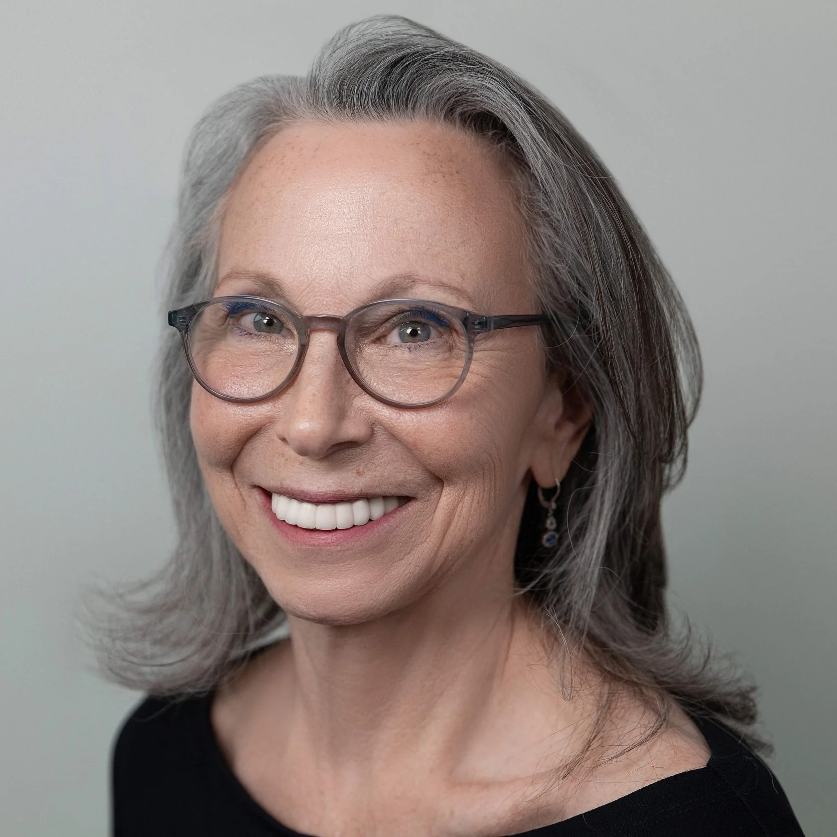 A headshot of a smiling middle-aged woman with gray hair, glasses, and earrings, wearing a black top against a plain background.