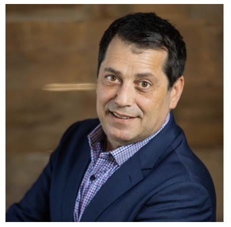 A professional man with dark hair wearing a blue blazer and dress shirt, smiling at the camera, with a wooden wall background.