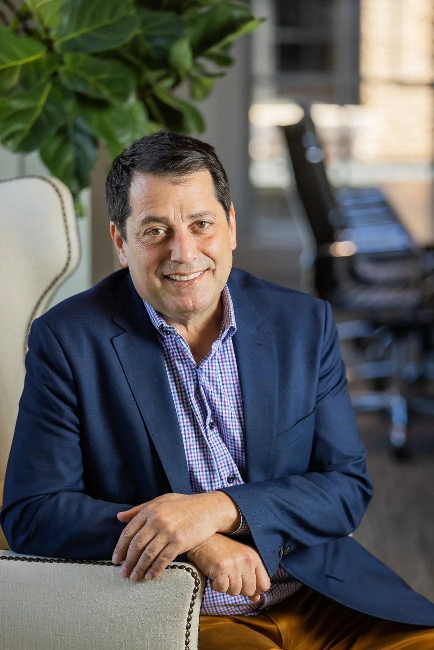 A man in a blue blazer and checkered shirt sitting at a desk in an office, smiling at the camera, with large green plants and office chairs in the background.