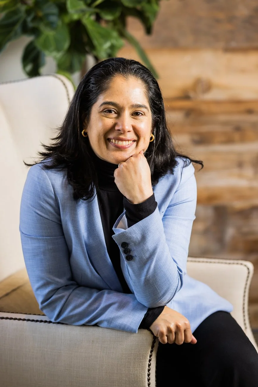 A woman with dark hair, wearing a light blue blazer and black turtleneck, sitting on a beige armchair and smiling at the camera with her chin resting on her hand. A leafy green plant and a wooden wall are visible in the background.