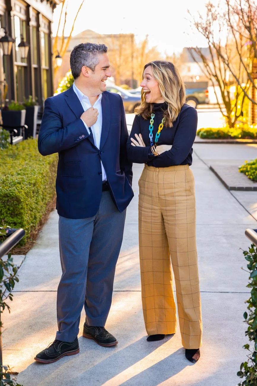 A man and woman standing outdoors on a sidewalk, laughing and talking to each other, with greenery and parked cars in the background, during sunset.