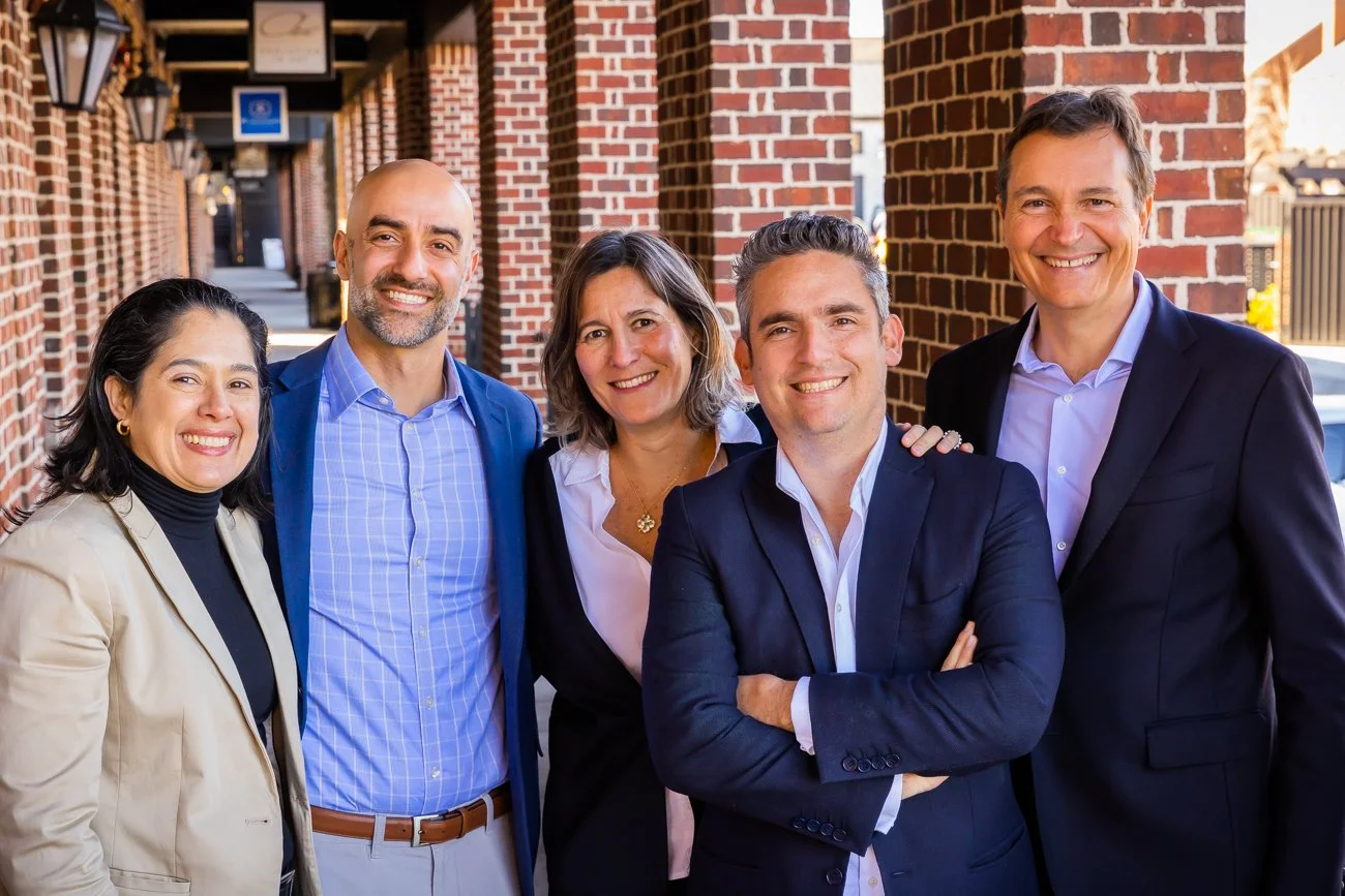 Group of five diverse professionally dressed people smiling outdoors against a brick building.