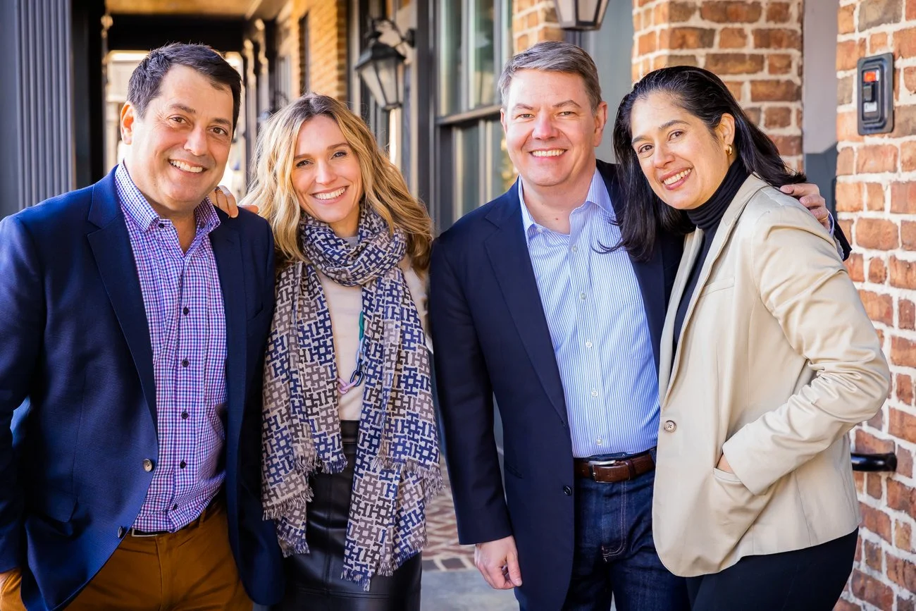 Group of five professionally dressed adults smiling and standing close together outside against a brick wall.
