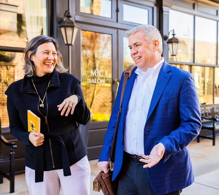 A man and woman are standing outside in conversation, both smiling and laughing. The woman holds a yellow notebook, and the man carries a brown shoulder bag. Behind them is a building with large glass windows and a sign that reads 'MIC SALON.'