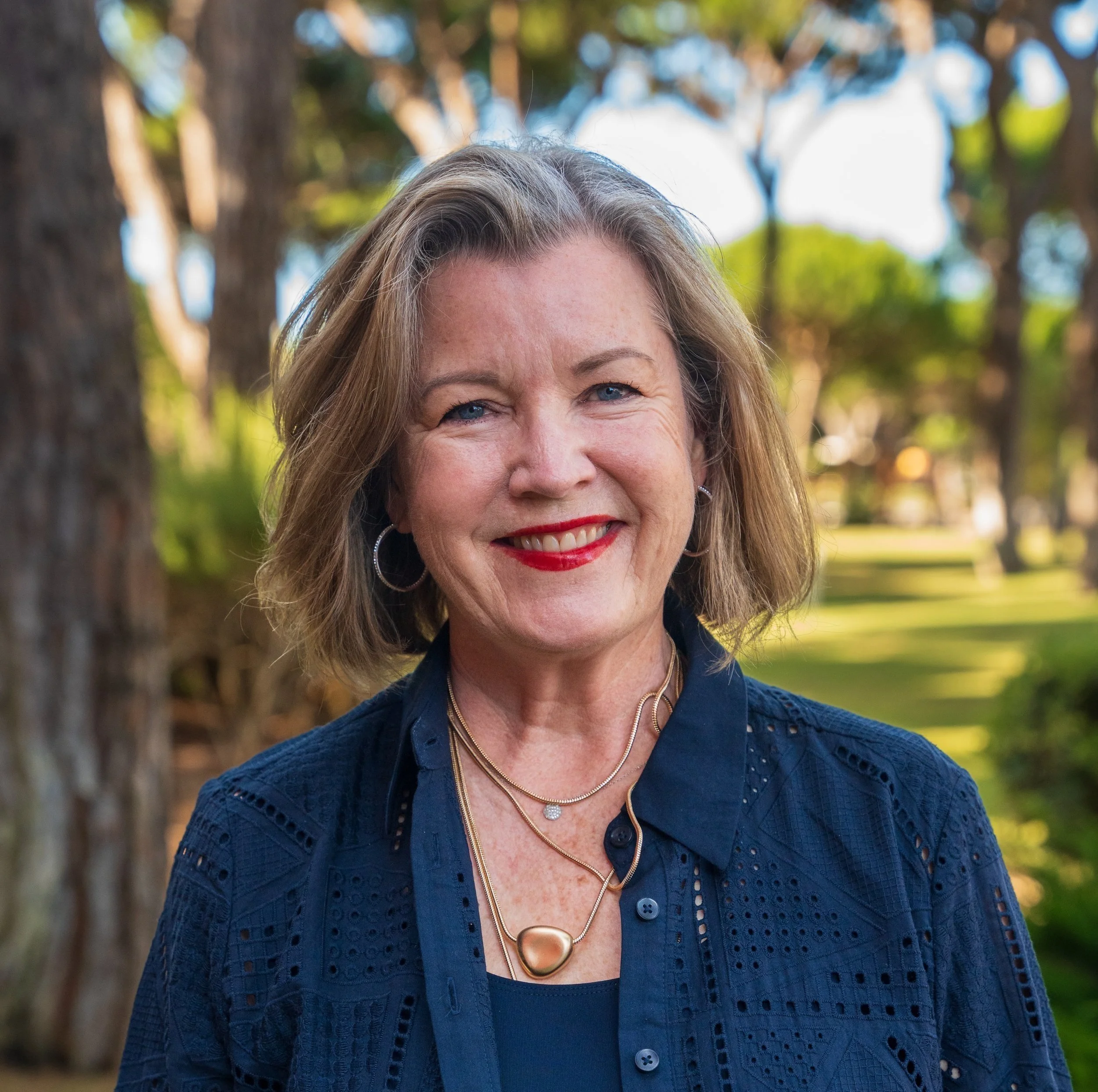 A woman smiling outdoors in a park with trees in the background, wearing a navy blue top and gold jewelry.