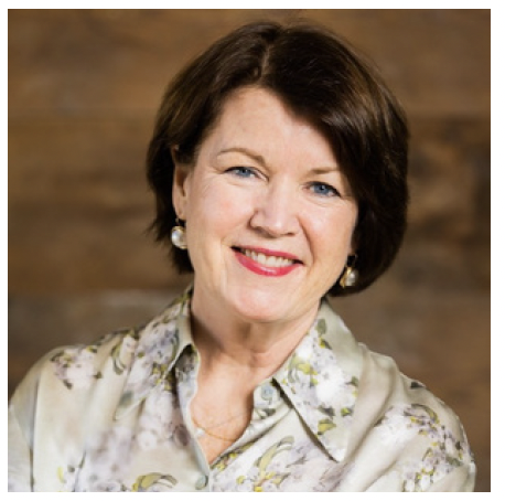 Portrait of a smiling older woman with short dark brown hair, wearing a floral blouse and earrings, against a wooden background.