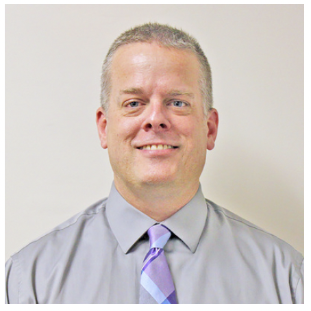 A man with short gray hair, light skin, smiling, wearing a light gray dress shirt and a purple striped tie, standing against a plain white background.