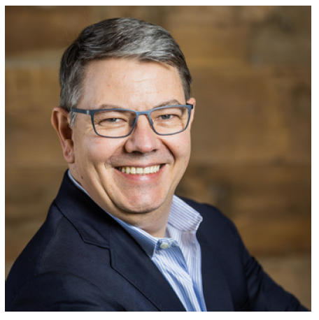 Portrait of a smiling man with glasses, wearing a dark blazer and light blue shirt, in front of a wooden background.