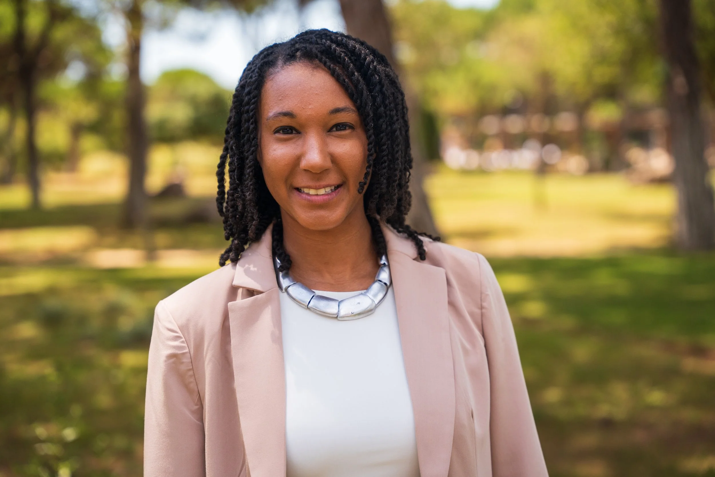 A woman with dark skin and black curly hair standing outdoors in a park with trees and grass in the background. She is wearing a beige blazer, white top, and a silver necklace, smiling at the camera.