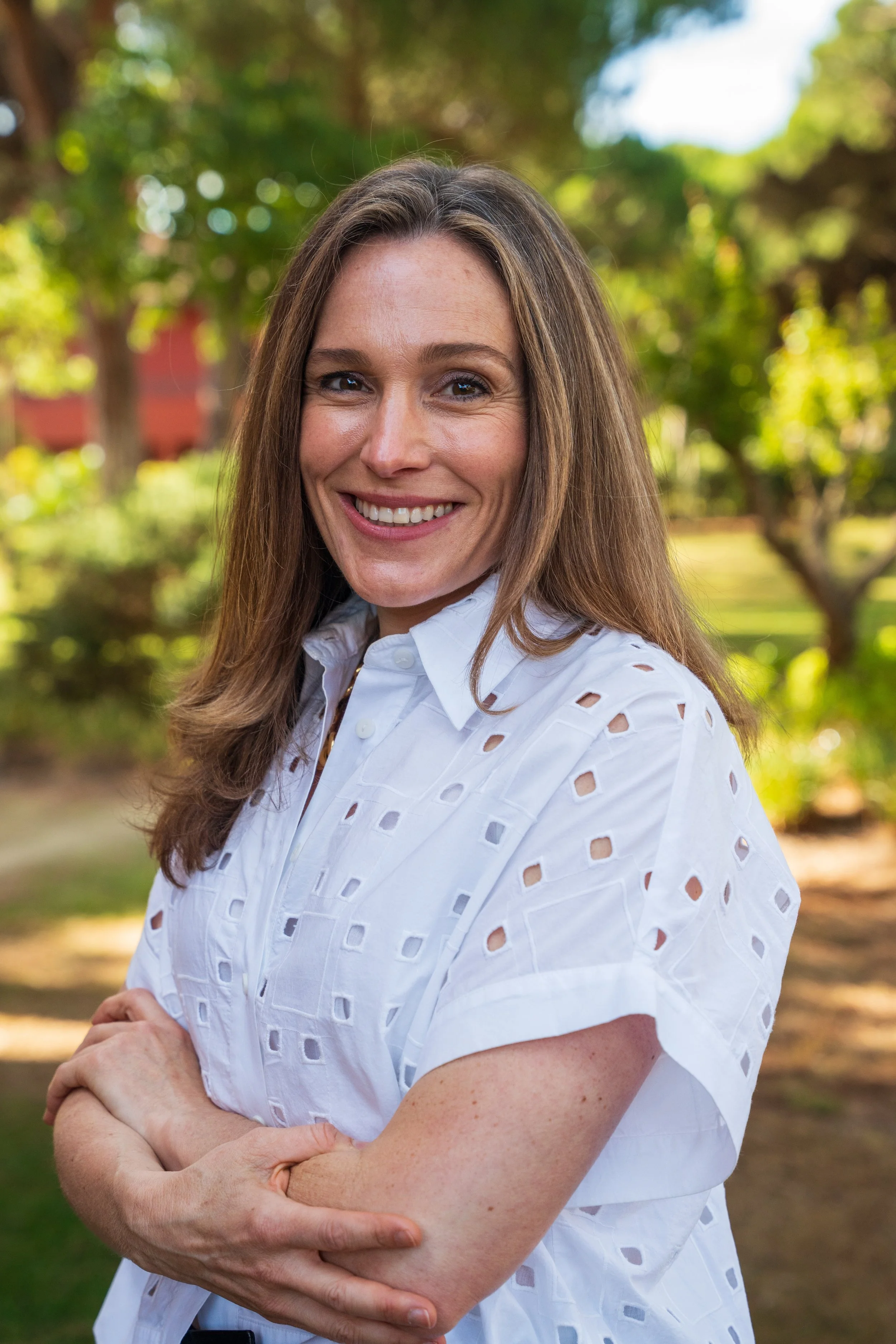 A smiling woman with long brown hair wearing a white perforated blouse, standing outdoors with trees and greenery in the background.