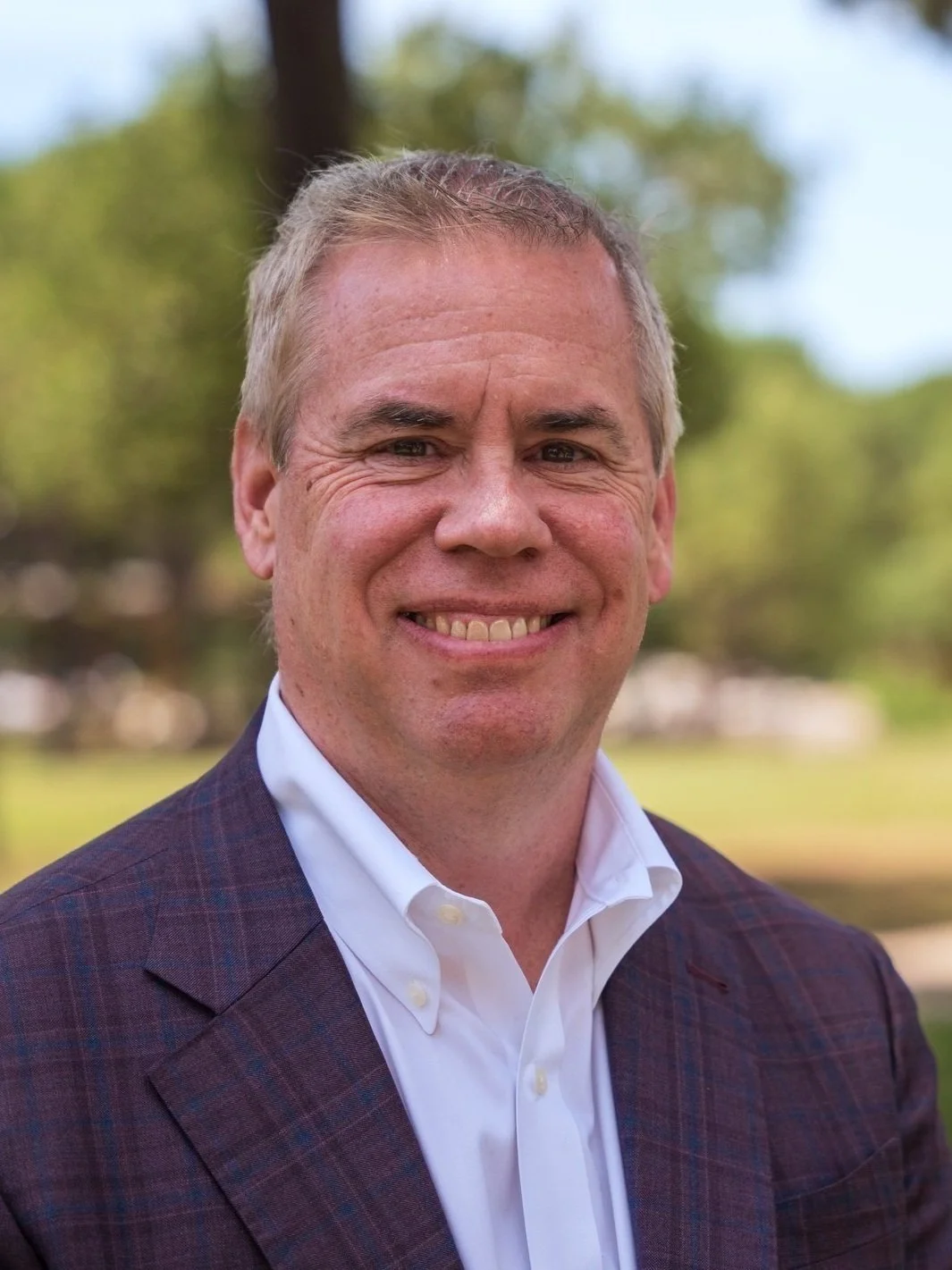 A smiling man with short blonde hair wearing a dark blazer and white shirt outdoors with trees and a park in the background.