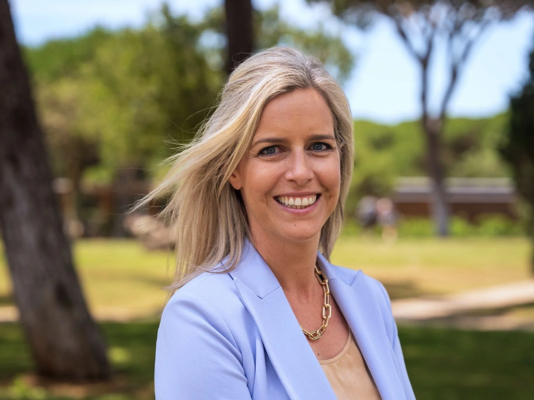 Headshot of a woman smiling outdoors, wearing a light blue blazer and a gold chain necklace, with trees and a park in the background.